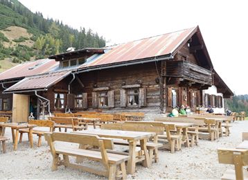 Une maison en bois traditionnelle dans les montagnes avec une terrasse et plusieurs tables en bois. À l'arrière-plan, on peut voir des collines boisées et un paysage naturel.