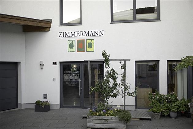 A modern building with the inscription "ZIMMERMANN" above the entrance.  
In front of the entrance, there are green plants and flowers in pots.