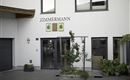 A modern building with the inscription "ZIMMERMANN" above the entrance.  
In front of the entrance, there are green plants and flowers in pots.