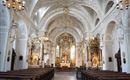 An elegant church with a high, intricately decorated vault. The altar area is clearly visible and surrounded by wooden pews.