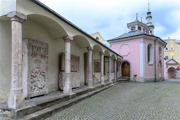 A historic architecture with columns and reliefs. In the background stands a pink chapel under a cloudy sky.