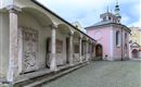A historic architecture with columns and reliefs. In the background stands a pink chapel under a cloudy sky.