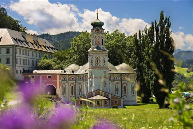 Eine malerische Kirche umgeben von grünen Wäldern und blühenden Blumen. Der Himmel ist blau mit wenigen Wolken.