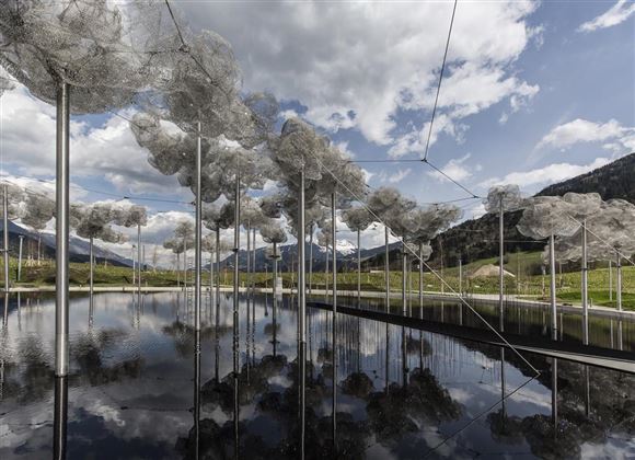 Un parco d'arte moderno con nuvole in materiale a rete che fluttuano sopra un'acqua calma. Sullo sfondo si vedono montagne e un cielo blu.
