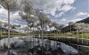 Un parco d'arte moderno con nuvole in materiale a rete che fluttuano sopra un'acqua calma. Sullo sfondo si vedono montagne e un cielo blu.
