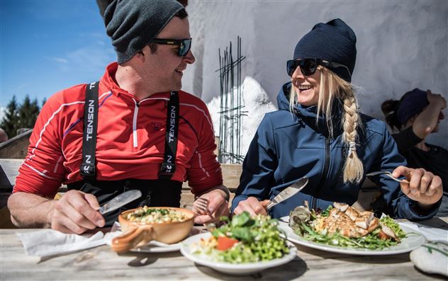 Zwei Personen genießen ein gemütliches Essen im Freien, während sie in Winterbekleidung gekleidet sind. Auf dem Tisch liegen zwei Teller mit Salat und einem herzhaften Gericht.
