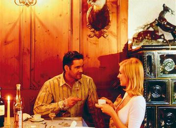 Un restaurant cosy avec des lambris en bois, où un homme et une femme parlent à la lumière des bougies. En arrière-plan, on voit de belles décorations et une cheminée.