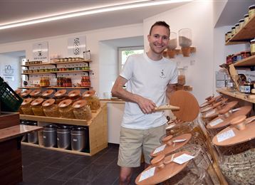 Een vriendelijke verkoper staat in een milieuvriendelijke winkel vol voorraadpotten met voedsel. Op de achtergrond zijn planken met verschillende producten zichtbaar.