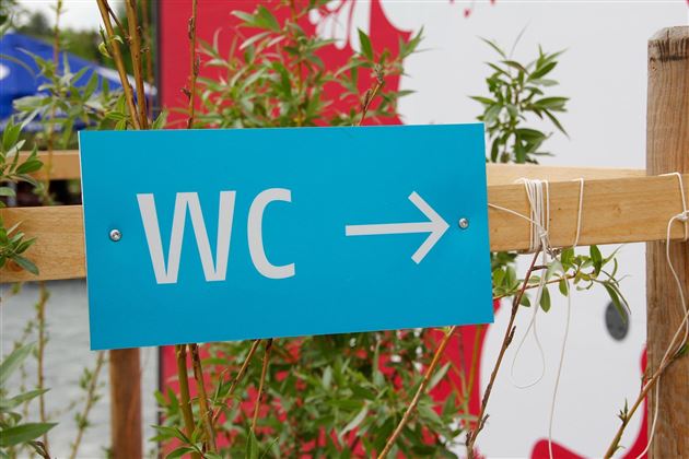 Een blauw bord wijst in de richting van het toilet. Het is omringd door planten.