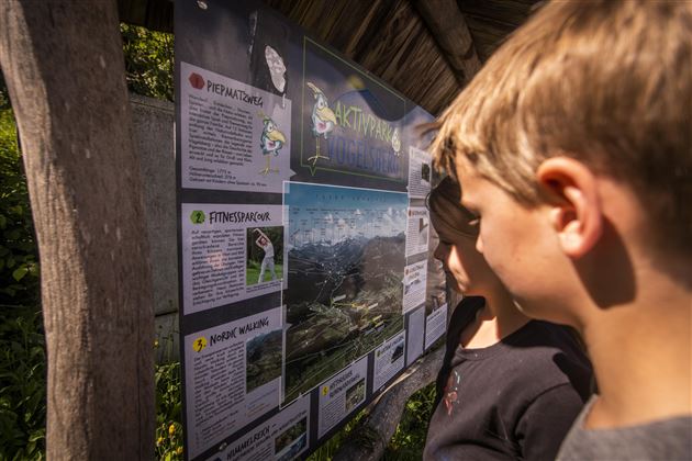 Zwei Kinder betrachten ein Informationsschild über die Natur. Das Schild enthält Bilder und Texte zu verschiedenen ökologischen Themen.