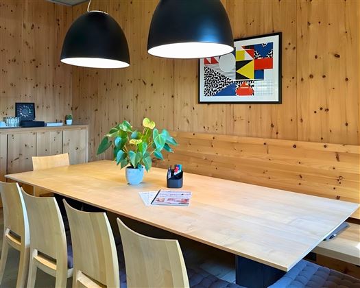 Ein moderner Besprechungsraum mit einem großen Holztisch und sechs Stühlen. An der Wand hängt ein farbenfrohes Kunstwerk und ein Pflanze steht auf dem Tisch.
