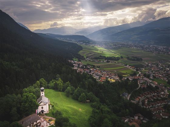 Een schilderachtig landschap met zachte heuvels en een klein dorp. Op de voorgrond staat een charmante kerk, omringd door weelderig groen en bergen op de achtergrond.
