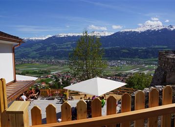 Una bella vista su un paesaggio vallivo verde con montagne coperte di neve sullo sfondo. Sulla terrazza, le persone godono del sole sotto un ombrellone.