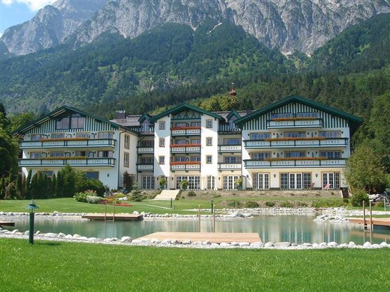 Ein charmantes Hotelgebäude mit mehreren Stockwerken, umgeben von grünen Wiesen und einem klaren Teich. Im Hintergrund erheben sich majestätische Berge unter einem blauen Himmel.