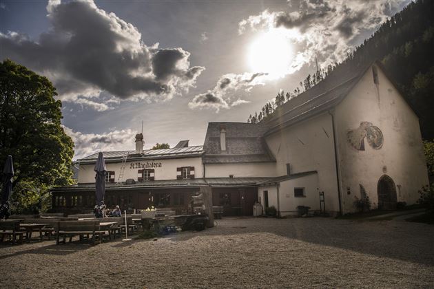 Een charmant gebouw met een terras buiten en bomen op de achtergrond. De lucht is bewolkt en de zon schijnt door de wolken.