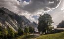 Een schilderachtig berglandschap met hoge bergen en een zachte groene weide. Op de achtergrond zijn er wolken en bomen te zien die een rustige sfeer creëren.