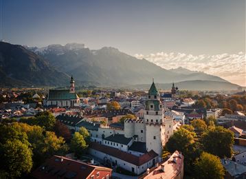 A picturesque cityscape with high mountains in the background. The image shows historic buildings and green foliage in the surroundings.