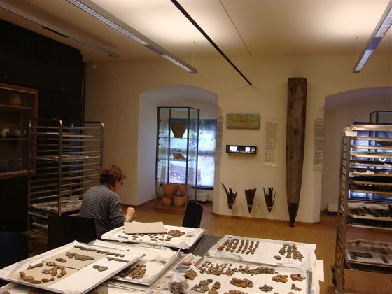 Ein Raum mit Tischen, die mit archäologischen Artefakten und Materialien zum Arbeiten bedeckt sind. Im Hintergrund sitzt eine Person und arbeitet an einem Projekt.