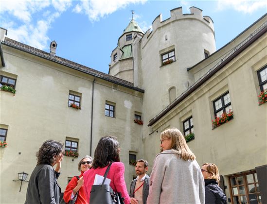 A group of people is standing in the courtyard of a historic castle. In the background, towers and windows with flowers are visible.