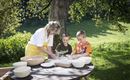 Eine Frau und zwei Kinder machen zusammen Teig im Freien. Der Tisch ist mit Schüsseln und Zutaten dekoriert, umgeben von einem grünen Garten.