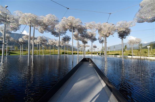 Een artistieke installatie met zwevende, wolkenachtige structuren boven een rustige waterpartij. Op de achtergrond zijn groene weiden en zachte heuvels zichtbaar.