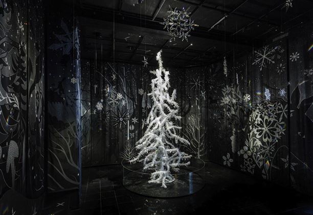Un albero di Natale bianco e luminoso si trova in una stanza buia con motivi murali creativi e grafici. Attorno all'albero sono appese decorazioni simili alla neve e fiocchi di neve.