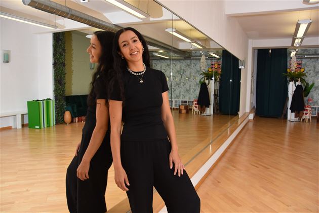 A woman stands in a dance studio in front of a mirror. The room has a light wooden floor and is warmly furnished.