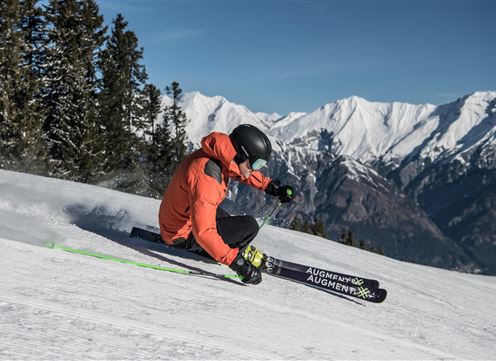 Ein Skifahrer in einem orangefarbenen Anzug fährt dynamisch einen schneebedeckten Hang hinunter. Im Hintergrund sind majestätische Berge und blauer Himmel zu sehen.
