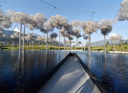 Eine künstlerische Installation mit schwebenden, wolkenartigen Strukturen über einem ruhigen Wasserbecken. Im Hintergrund sind grüne Wiesen und sanfte Hügel sichtbar.