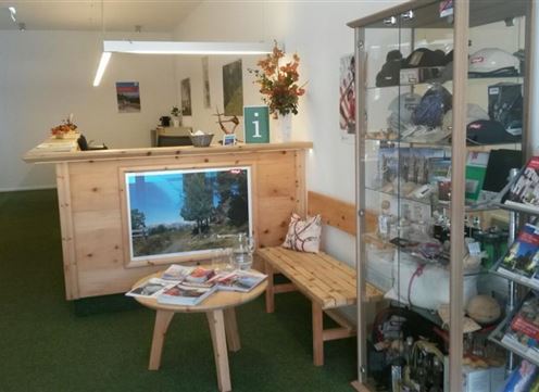 Un'area ricevimento con bancone in legno e materiale informativo. Su un tavolo ci sono opuscoli e un grande schermo mostra un bel paesaggio.