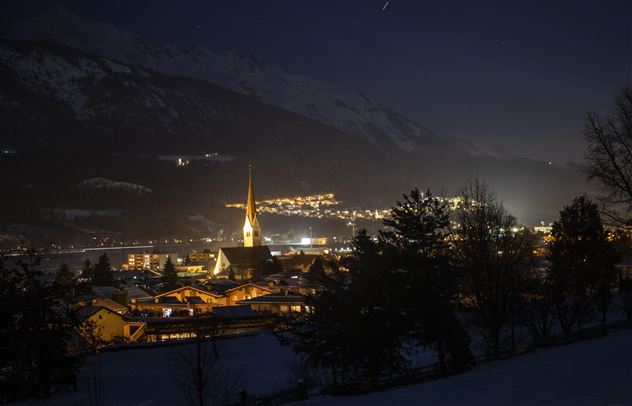Eine malerische Berglandschaft bei Nacht mit beleuchteten Gebäuden. Die Kirche mit dem hohen Turm strahlt im Licht der Stadt.