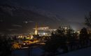 Eine malerische Berglandschaft bei Nacht mit beleuchteten Gebäuden. Die Kirche mit dem hohen Turm strahlt im Licht der Stadt.