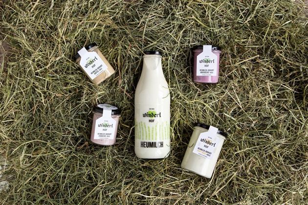 Eine Flasche Heumilch steht in der Mitte, umgeben von verschiedenen Joghurtbechern. Alle Produkte sind auf einem Bett aus Heu arrangiert.