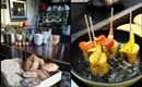 A cozy café with a counter full of drinks and fresh bread. On the other side, grilled corn on the cob with colorful vegetables can be seen.