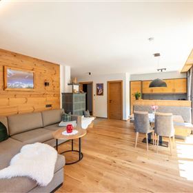 A stylish living and dining room with wooden paneling and large windows. The room is bright and modernly furnished, with a comfortable sofa and a dining table.