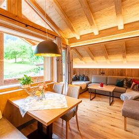 A cozy living room with wooden furniture and large windows. The light floods the room and offers a view of the green landscape outside.