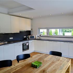 Een moderne keuken met witte kasten en een grote houten tafel. Door het raam valt veel licht binnen en er is een mooi uitzicht op het groen.