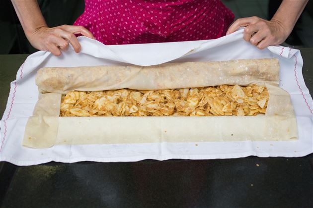 Ein Mensch rollt einen Teig mit einer Füllung aus Äpfeln und Gewürzen. Der Teig liegt auf einem weißen Tuch und wird sorgfältig vorbereitet.