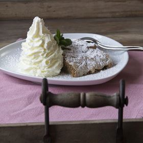Ein Stück Kuchen mit Puderzucker und eine große Portion Schlagsahne auf einem weißen Teller. Das Ganze ist auf einem Holztablett mit einem rosa Tuch angerichtet.