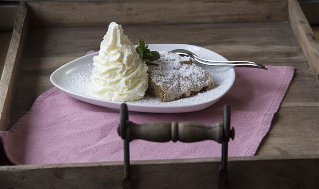 Ein Stück Kuchen mit Puderzucker und eine große Portion Schlagsahne auf einem weißen Teller. Das Ganze ist auf einem Holztablett mit einem rosa Tuch angerichtet.