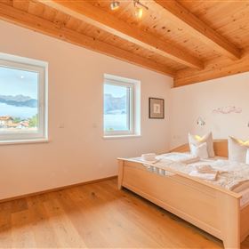 A modern bedroom with a wooden ceiling and large windows. The view extends over a picturesque landscape with mountains and water.