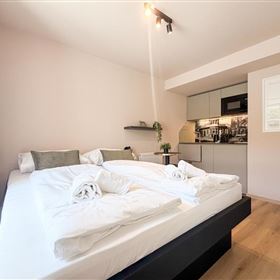 Ein modernes Schlafzimmer mit zwei Einzelbetten und frischen Handtüchern. Im Hintergrund befindet sich eine Küchenzeile und helle Farben sorgen für eine einladende Atmosphäre.