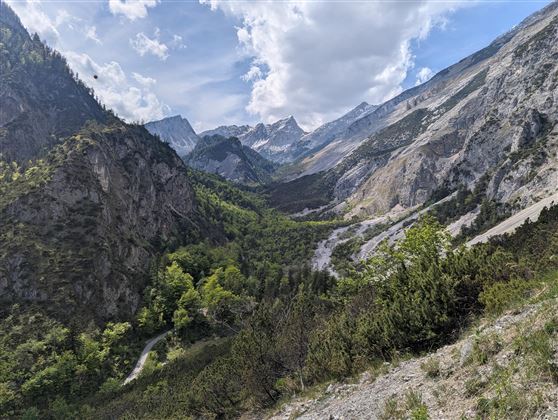 Un paesaggio montano pittoresco con alte montagne e una valle verde. Il cielo è parzialmente nuvoloso con alcuni raggi di sole.