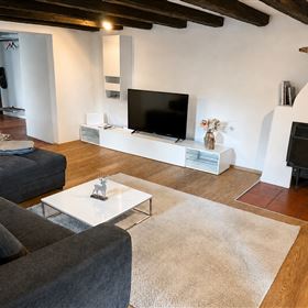 Ein modernes Wohnzimmer mit einem großen, bequemen Sofa und einem flachen Fernseher auf einem weißen TV-Möbel. Der Raum hat Holzdeckenbalken und einen gemütlichen Kamin.
