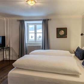 A cozy bedroom with two single beds, a television, and a window looking outside. The walls are light and decorated with a picture, while the curtains are in soft colors.