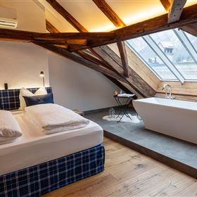 A modern room with a cozy bed and large windows. On the right side, there is a freestanding bathtub.