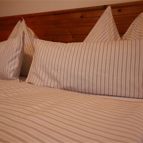 Ein gemütliches Bett mit gestreiften Kissen und einer weichen Decke. Der Holzwand im Hintergrund sorgt für eine warme Atmosphäre.