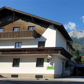 Ein modernes Gebäude mit Holzverkleidung und Balkonen in den Alpen. Im Hintergrund sind majestätische Berge und blauer Himmel zu sehen.