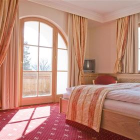 Una camera d'hotel accogliente con un grande letto e un tappeto rosso. Le finestre lasciano entrare molta luce diurna e offrono una vista verso l'esterno.