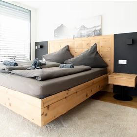 Een moderne slaapkamer met een groot houten bed en grijze beddengoed. Naast het bed staat een nachtzwart tafeltje en grote ramen laten veel licht binnen.